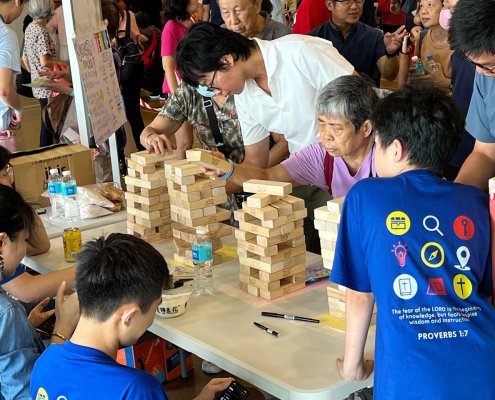 Multiple kids and adults crowd around a Giant Jenga game in Singapore, sharing smiles and playful competition.