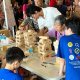 Multiple kids and adults crowd around a Giant Jenga game in Singapore, sharing smiles and playful competition.