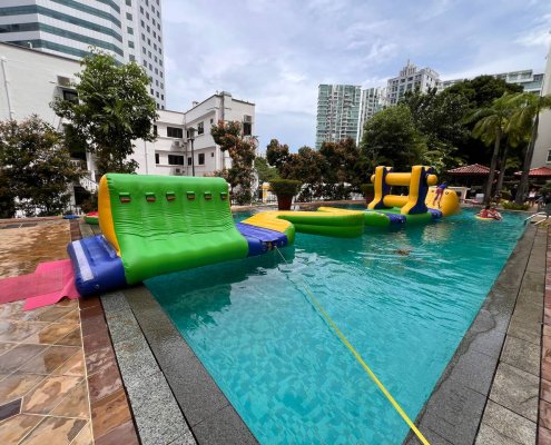 Inflatable Cliff obstacle in green and yellow with blue climbing handles, shaped like a triangle, floating on water.