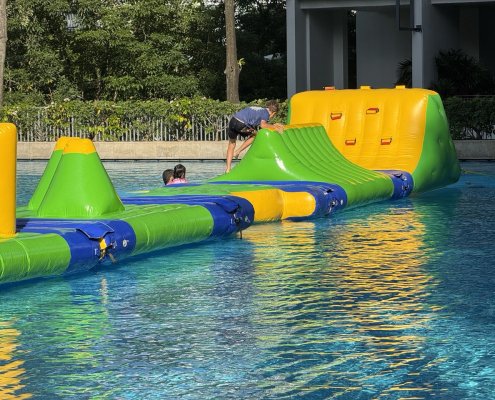 Participant climbing the inflatable Cliff obstacle using blue handles, part of a colorful water obstacle course.