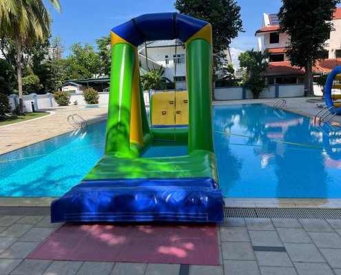 A triangular green inflatable structure with a rope hanging in the middle, part of a water obstacle course designed for swinging across the water.