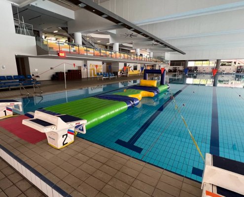 A yellow inflatable square platform next to a blue inflatable square with a low opening underneath, part of a water obstacle course.