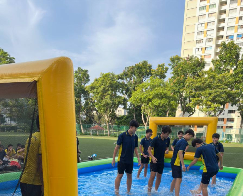 Water Soccer -Bishan Street Students at Bishan Street, Singapore laughing and kicking a ball on a bright blue and yellow inflatable water soccer field, splashing water as they play.