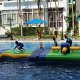 Kids walking across The Wave, a flat green inflatable platform with wavy edges in a water obstacle course.