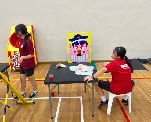 A colorful character-themed game board showing a wide-mouthed face with teeth designed to pop out when hit by soft balls — set up at MOE Evans Stadium, Singapore.