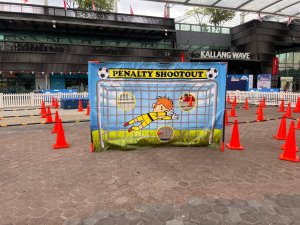 The Penalty Shootout game shows a printed goalkeeper on a banner with three cut-out holes as targets, inviting players to test their football skills at OCBC Square, Singapore