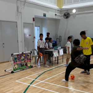 A student kicks a football toward a goal covered with a banner featuring five cutouts, aiming to score through the holes during a game session at Shuqun Primary School, Singapore.