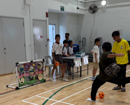 A student kicks a football toward a goal covered with a banner featuring five cutouts, aiming to score through the holes during a game session at Shuqun Primary School, Singapore.